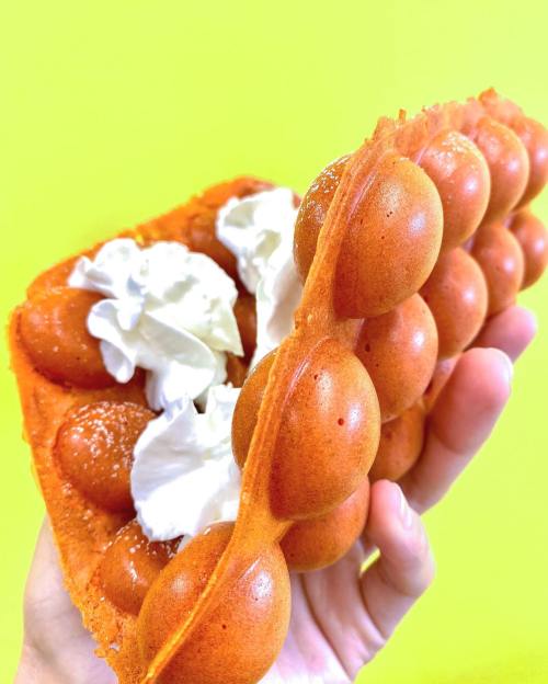 A hand holding a golden-orange bubble waffle filled with whipped cream against a bright yellow background.