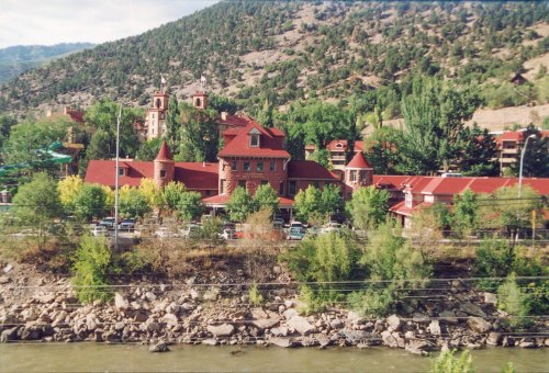 Historic red-roofed building surrounded by trees and mountains, with a river in the foreground.