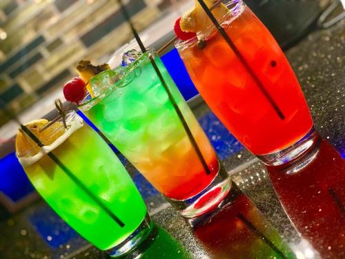 Three colorful cocktails in tall glasses: green, orange, and red, garnished with fruit and ice, on a reflective surface.