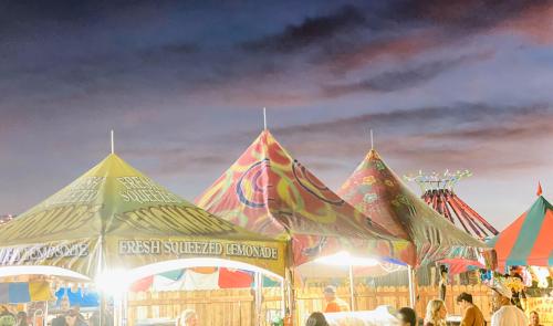 Colorful tents at a fair serve fresh squeezed lemonade under a twilight sky, with carnival rides in the background.