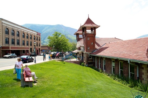 A sunny street scene with people walking, a historic building, and mountains in the background. Green grass in the foreground.