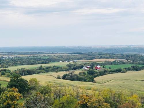 A scenic view of rolling green hills with a few red barns and trees under a cloudy sky.