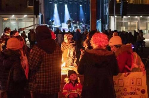 A crowd gathers around a fire at an outdoor event, with a stage and performers visible in the background.