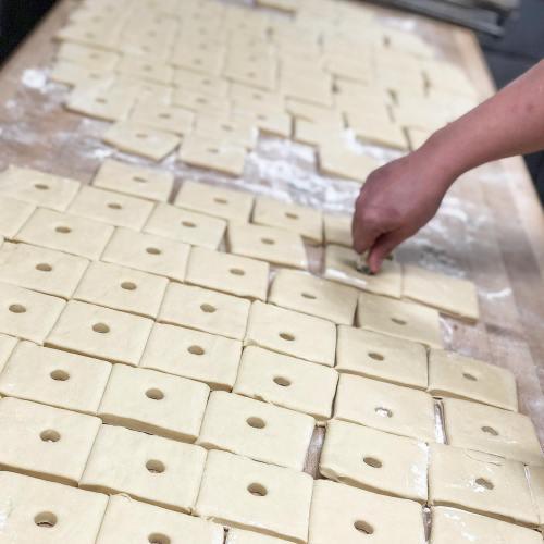 A hand places a small piece of dough on a table covered with neatly arranged rectangular dough pieces with holes.