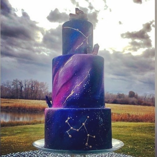 A three-tiered galaxy-themed cake with purple and blue swirls, adorned with crystals and constellations, set outdoors.