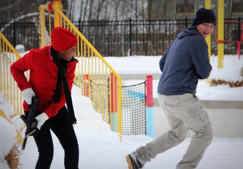 A person in a red jacket holds a toy gun while another person in a blue jacket runs in a snowy playground.