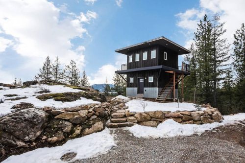 A modern black cabin on a rocky landscape, surrounded by snow and pine trees under a cloudy sky.