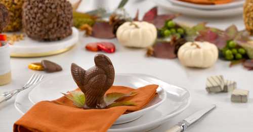 A festive table setting with a chocolate turkey on an orange napkin, surrounded by autumn leaves and decorative pumpkins.