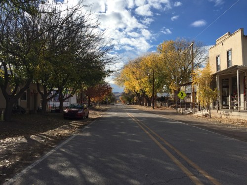 A quiet street lined with trees displaying autumn colors, with a few buildings and a parked car on the side.