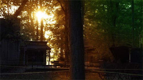 Sunlight filters through trees, illuminating a rustic scene with buildings and a vintage wagon in a serene setting.