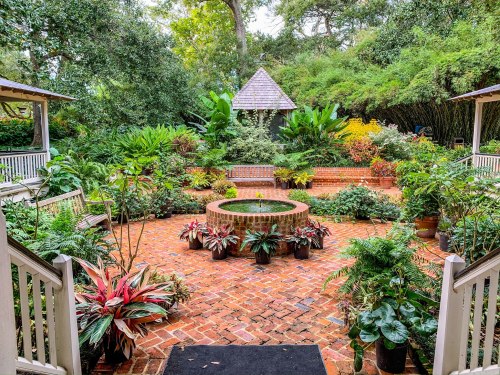 A lush garden with a circular brick fountain, surrounded by vibrant plants and greenery, viewed from a staircase.