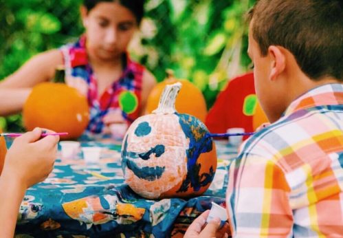 Children painting pumpkins at a table outdoors, surrounded by greenery and colorful decorations.