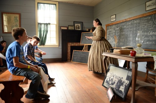 A teacher in period dress instructs students in a historic classroom with chalkboards and wooden benches.
