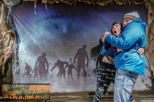 Two people pose in front of a spooky backdrop with zombie silhouettes, one looking scared and the other holding them tightly.
