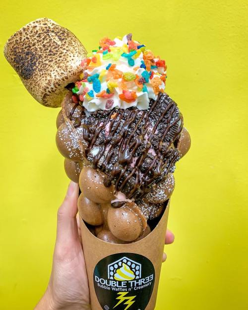 A hand holds a bubble waffle cone topped with chocolate, whipped cream, colorful sprinkles, and a toasted marshmallow.