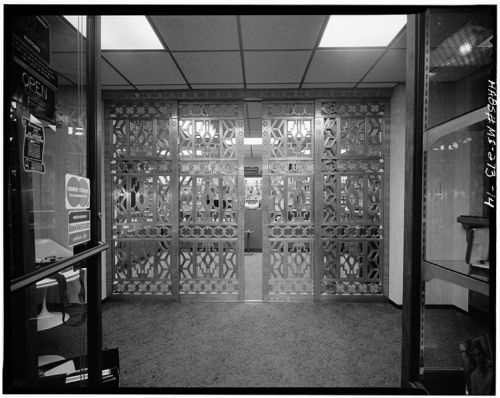 Black and white image of ornate metal gates leading into a building, with a carpeted entrance and glass doors.