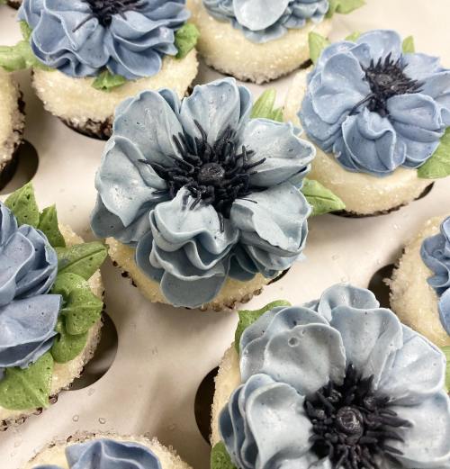 Close-up of cupcakes topped with blue flower-shaped frosting and green leaves, arranged in a tray.