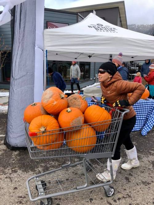A person pushes a shopping cart filled with large pumpkins at an outdoor market under a tent.