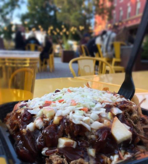 A close-up of a dish topped with coleslaw, served outdoors with blurred people and yellow chairs in the background.