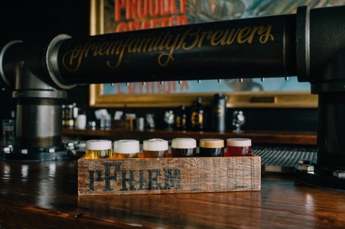 A wooden flight paddle holds six small glasses of different craft beers on a bar counter.