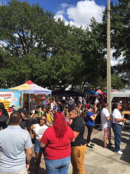 A bustling outdoor market scene with people mingling, food stalls, and trees under a blue sky.