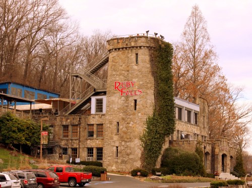 Historic stone building with "Ruby Falls" sign, surrounded by trees and parked cars in the foreground.
