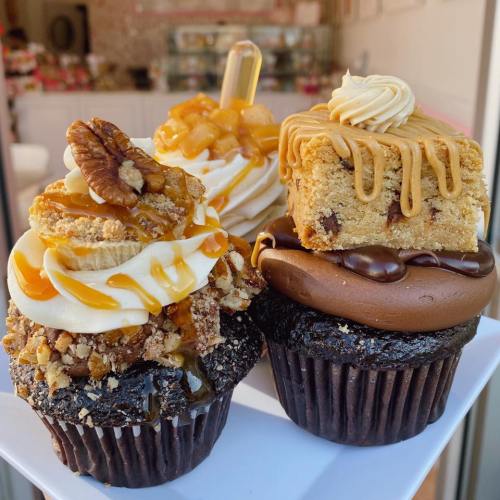 A close-up of four decadent cupcakes topped with various sweets, including caramel, nuts, and cookie pieces.