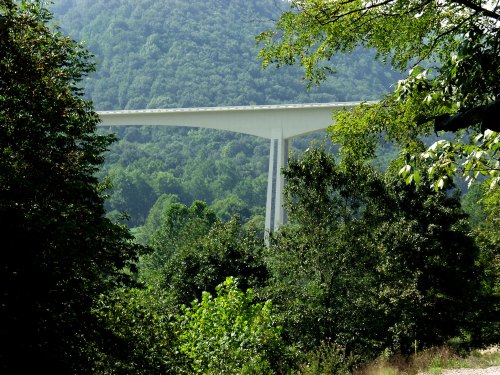 A modern white bridge spans a lush green valley, surrounded by dense trees and rolling hills in the background.