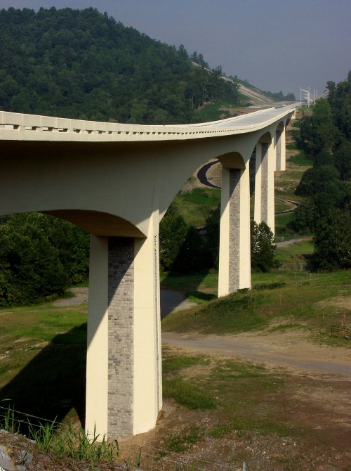 A modern bridge supported by tall pillars, spanning a green landscape with hills in the background.