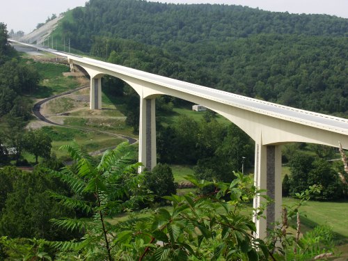A modern bridge spans a green valley, surrounded by trees and hills under a clear sky.