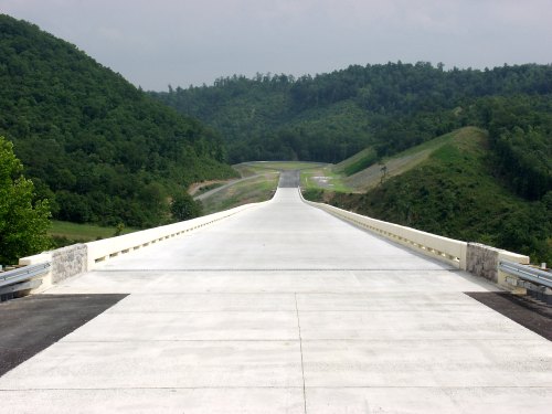 A wide, empty road stretches into the distance, flanked by green hills under a cloudy sky.