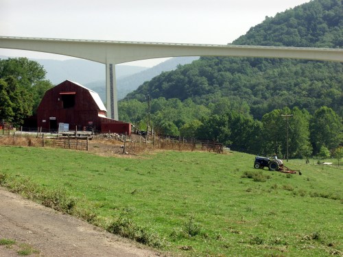 A red barn sits in a green field with a tractor nearby, under a large bridge and surrounded by rolling hills.