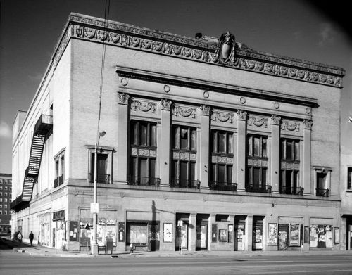 Historic theater building with ornate architectural details, featuring large windows and decorative friezes. Black and white photo.