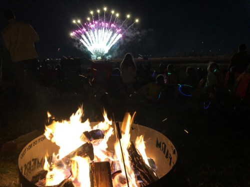 A campfire in the foreground with people watching colorful fireworks in the night sky.