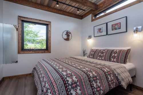 Cozy bedroom with a patterned bedspread, wooden beams, and a window overlooking greenery. Decorated with framed art.