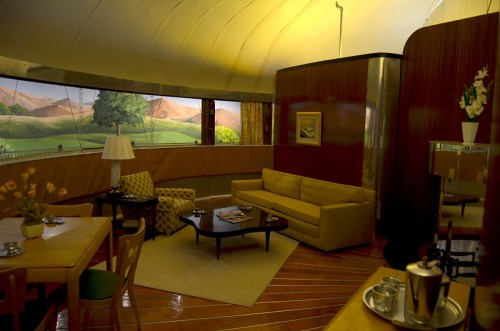 A cozy, retro living room with a yellow hue, featuring a sofa, dining table, and scenic wall mural of mountains.