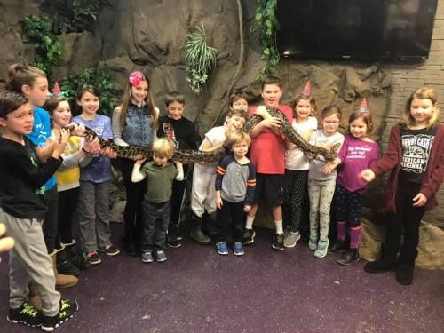 A group of children and adults pose together, smiling while holding a large snake in a decorated indoor setting.