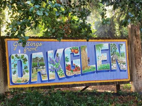 Colorful mural sign reading "Greetings from Oakglen," featuring fruits and flowers, surrounded by greenery.