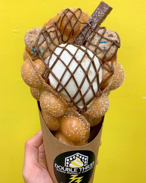 A hand holding a bubble waffle cone filled with ice cream, chocolate, and toppings against a bright yellow background.