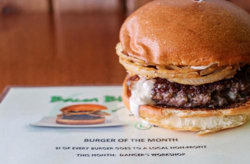Close-up of a burger with crispy onions on a bun, next to a sign promoting the burger of the month for a local non-profit.