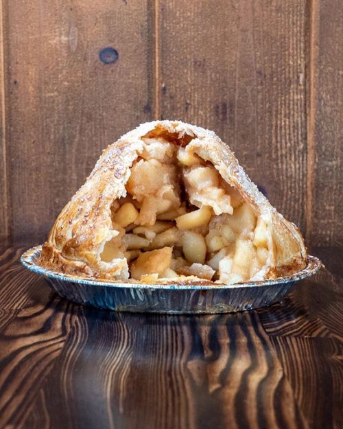 A golden-brown apple pie with a flaky crust, filled with sliced apples, sits in a silver pie dish on a wooden surface.