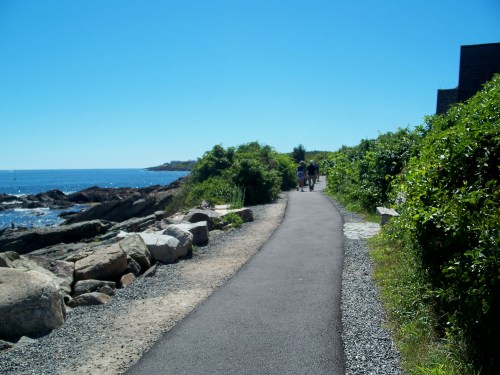 A scenic coastal path lined with greenery, leading to the ocean under a clear blue sky. People walk in the distance.