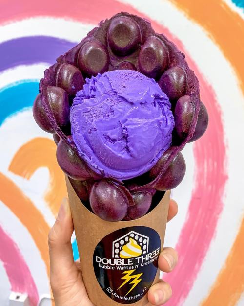 A hand holding a bubble waffle cone filled with purple ice cream against a colorful background.