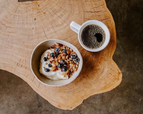 A bowl of yogurt topped with granola and blueberries, alongside a cup of coffee, on a wooden table.