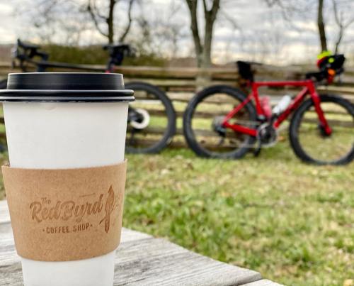 A coffee cup sits on a wooden table, with bicycles in the background and trees under a cloudy sky.