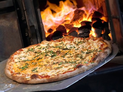 A freshly baked pizza on a metal peel, with a wood-fired oven and flames in the background.