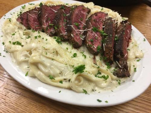 A plate of fettuccine Alfredo topped with sliced steak and garnished with parsley and grated cheese.