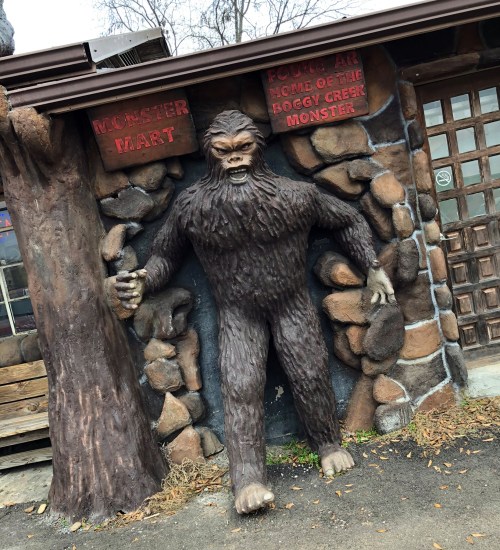 A large Bigfoot statue stands at the entrance of a shop, surrounded by a stone wall and a tree. Signs read "Monster Mart."