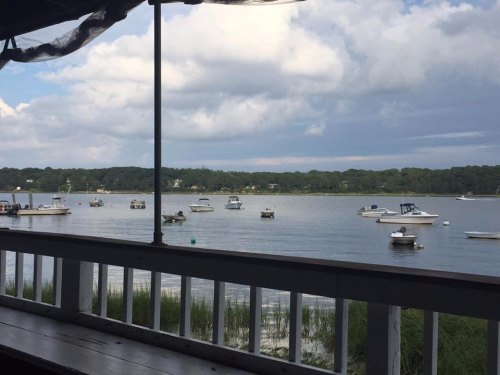 A serene view of a calm bay with several boats anchored, framed by a wooden railing and lush greenery in the background.