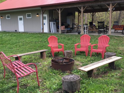 A cozy outdoor seating area with red chairs around a fire pit, surrounded by greenery and a rustic building in the background.
